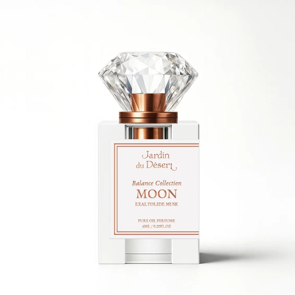 A perfume bottle from Jardin du Désert Balance Collection Moon Extrait de Rose, featuring a clear faceted diamond-shaped cap, a metallic rose gold collar, and a white rectangular bottle with a white label outlined in rose gold.